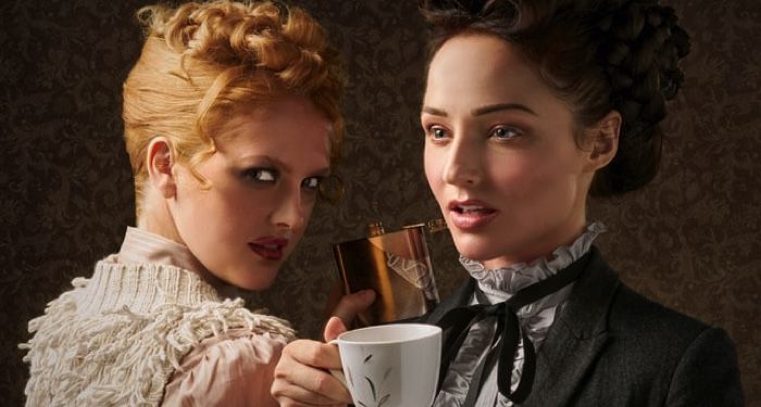two light-skinned women dressed in victorian clothing; one has a tea cup, the other is looking at the camera mysteriously
