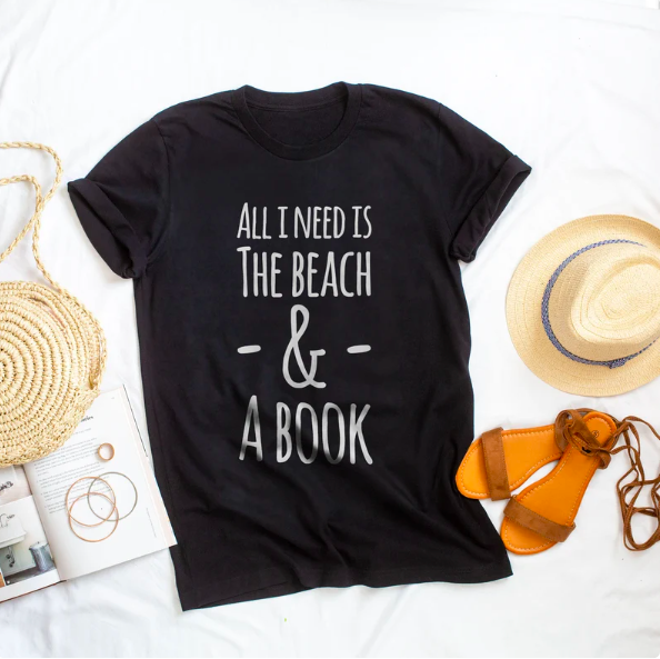 Black t-shirt that says, "All I need is the beach and a book" in white font, next to a sun hat and sandals