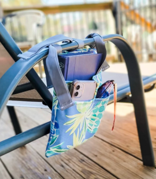Fabric caddy in blue floral print with pockets holding phone, book, and sunglasses, tied to the arm of a beach chair