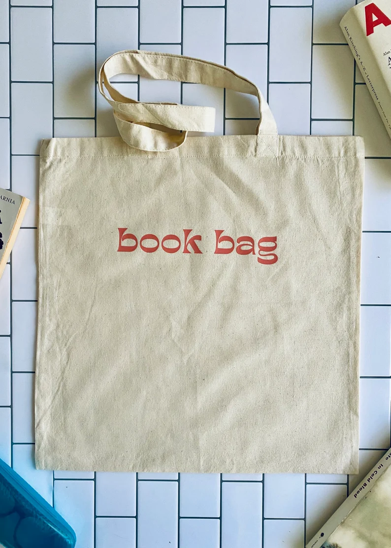 Image of a canvas tote on a tile background. In orange font it reads 
