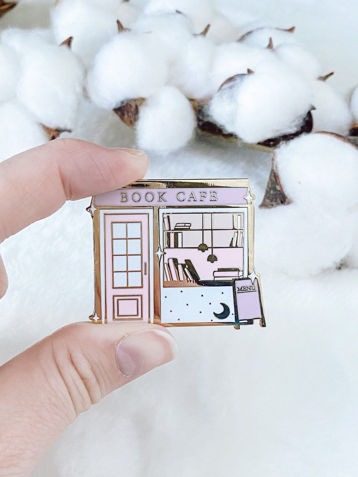 Image of a white hand holding an enamel pin. The pin is a pastel colored bookshop/cafe. 