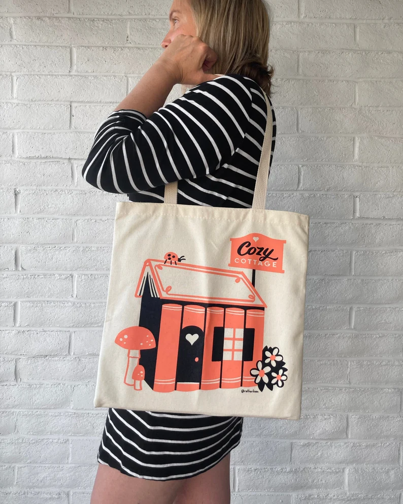 Image of a white person in a black and white striped dress. She has a canvas tote on her shoulder that features a house made of books. It's in coral and black, and it includes a sign reading 