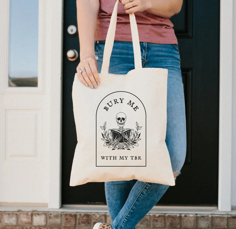 Image of a white person holding a canvas tote in front of a black door. The tote, which has black text, reads 