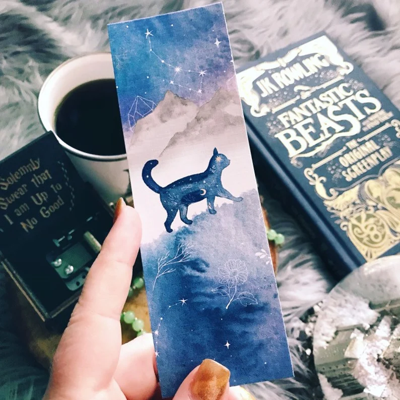 Image of a bookmark held by a white hand. the bookmark features a watercolor image including a cat with a constellation inside. 