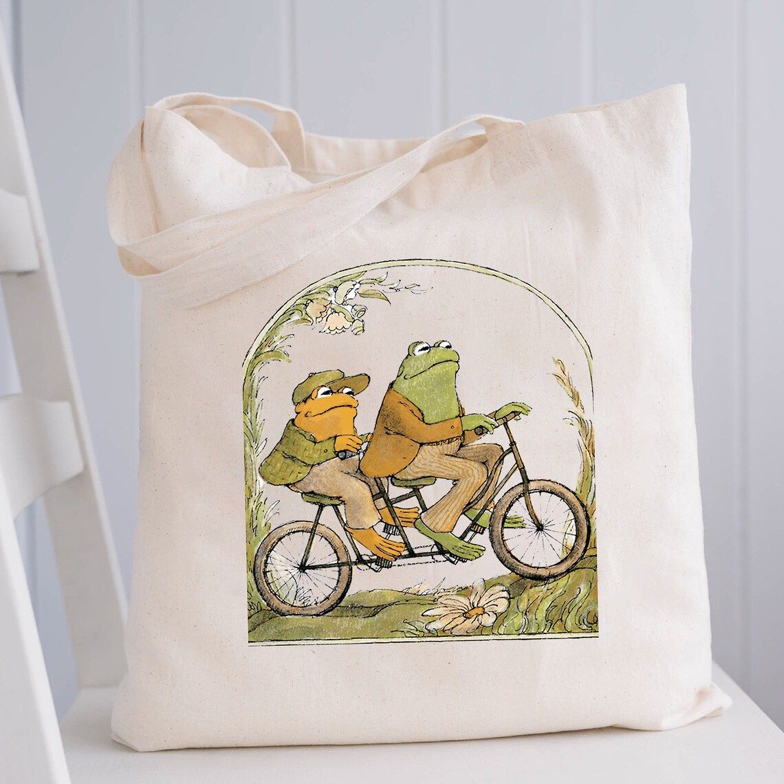 Beyaz bir sandalyede bir tuval çantası görüntüsü.  Bez çantanın üzerinde klasik Frog and Toad kitaplarından bir görüntü var.