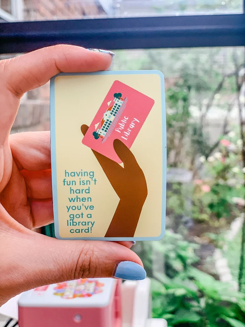 A hand holds a magnet. The magnet has a pastel yellow background. There is a black hand on it with a pink public library card and blue text reads "having fun isn't hard when you've got a library card."