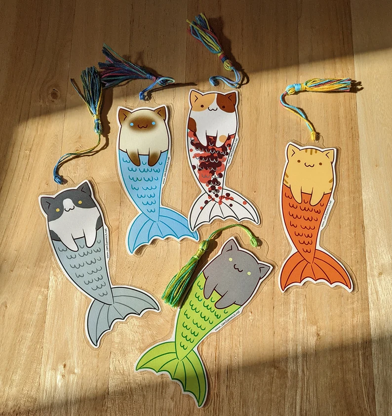 Image of five bookmarks on a wood background.. The cats each have a a mermaid tail. 