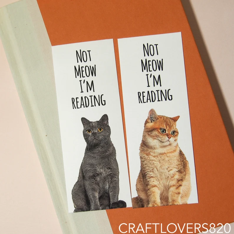 Image of two bookmarks on a book. The bookmarks both say "not meow, i'm reading." The bookmark on the left has a grey cat on the bottom and the bookmark on the right, an orange tabby cat. 