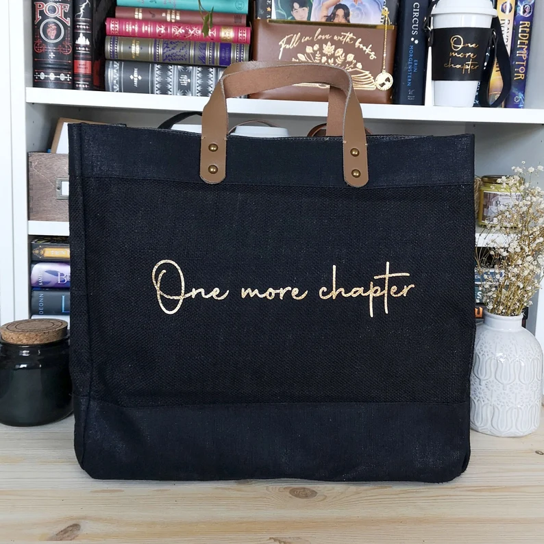 Image of a black tote bag with brown handles. There is cream script style text. It reads 