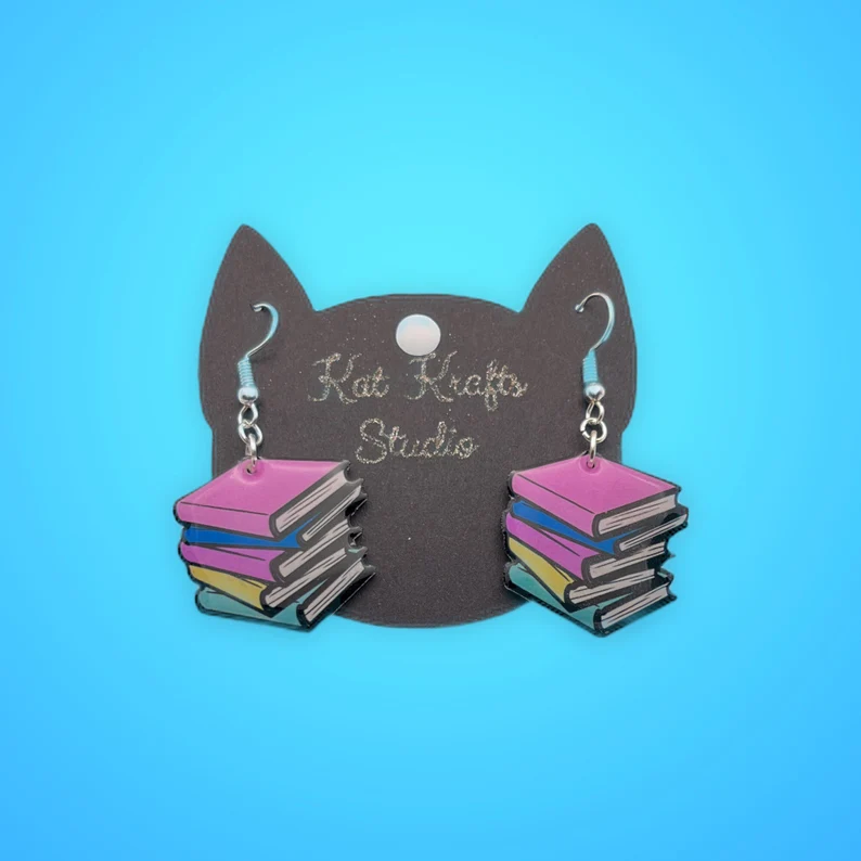 Image of a set of pastel earrings in the shape of book stacks. They are on a cat shaped display card on a blue background. 