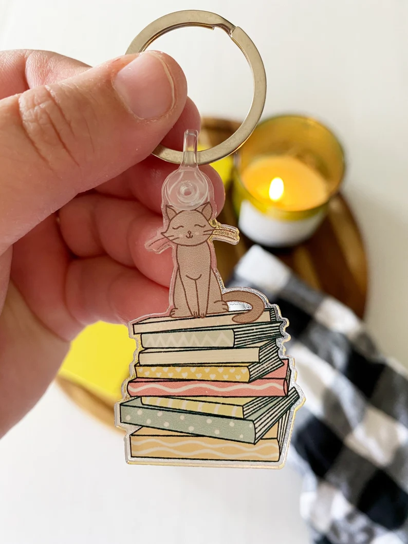 Image of a white hand holding a keychain. The keychain features a cat on a stack of pastel colored books. 