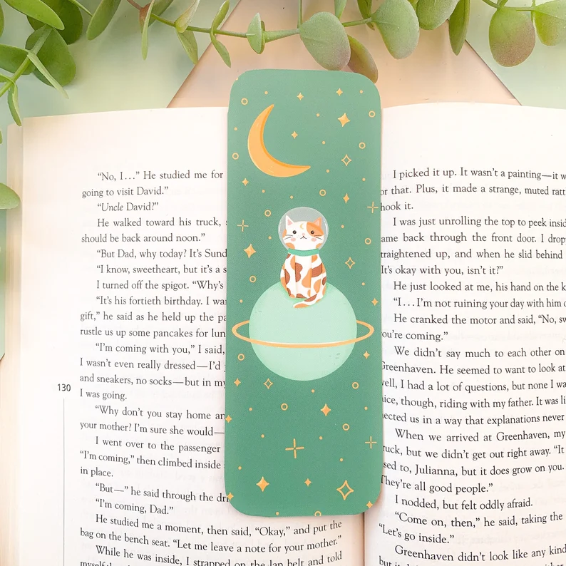 Image of a green bookmark on an open book. The bookmark has an orange spotted cat on top of a planet. The cat has on a space helmet. 