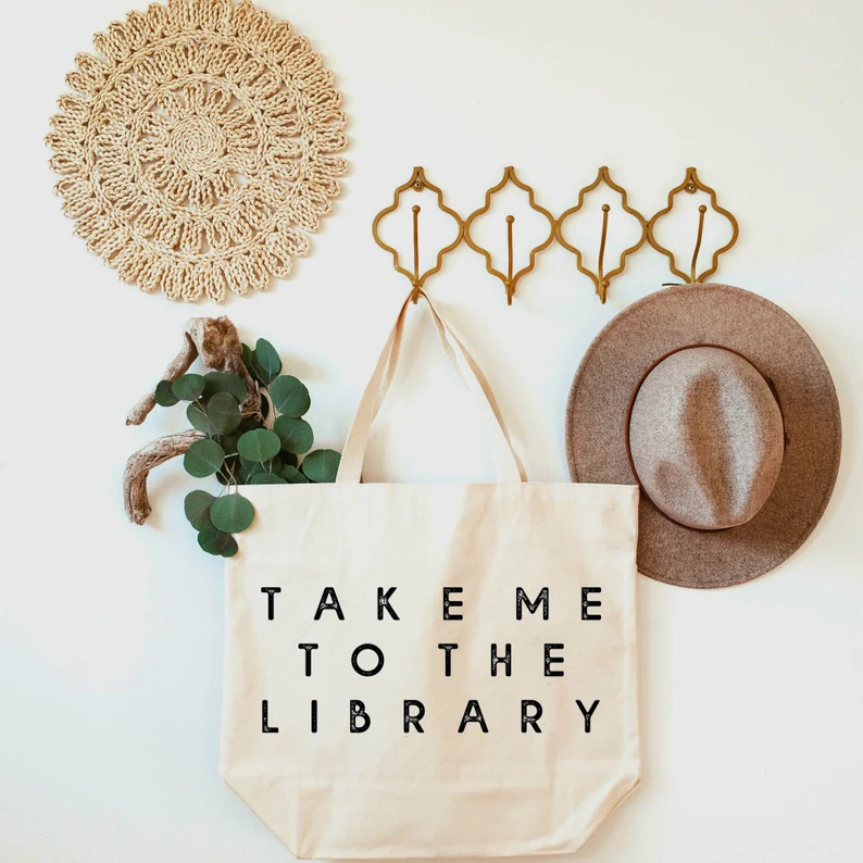Image of a large tote bag on a coat hook beside a hat and eucalyptus branch. The tote reads, in black font, 