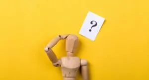 a small wooden mannequin with a question mark above it and one hand held up to its head as is in confusion or wonder.
