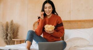 light-skinned Asian woman watching something on her laptop and smiling while eating from a bowl of popcorn