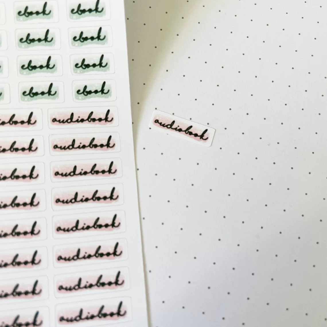 Image of a small sticker that reads "audiobook" in script stuck on a dotted journal page. To the left is a large sticker sheet with several of the audiobook stickers, and others that read "ebook."