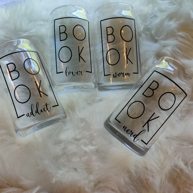 Four glass tumblers on a white furry background. The mugs have black writing. The writing says "book addict," "book lover," "book nerd," and "book worm."