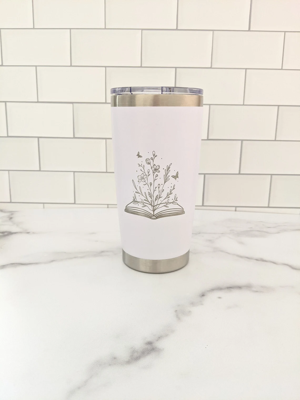 Image of a white tumbler on a marble counter with white tiles on the wall behind it. The tumbler features a black line drawing of an open book with flowers and insects. 