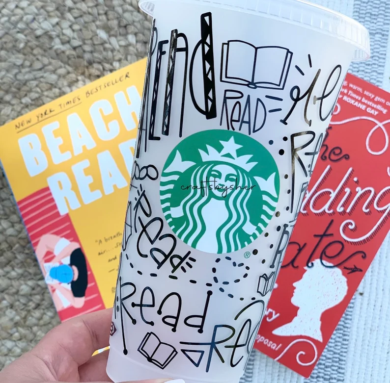 Image of a clear starbucks reusable cup. It's been customized with words like "read" and with images of books. It is in front of two books being held by a light colored hand. 