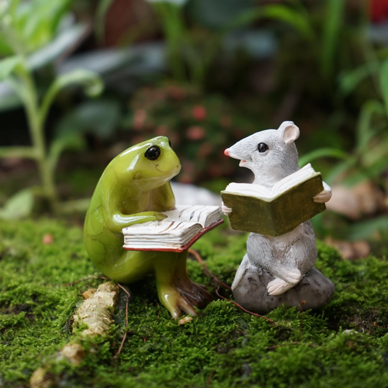 Two tiny statues of a green frog and a white mouse, both holding open books, sit on a bed of moss.