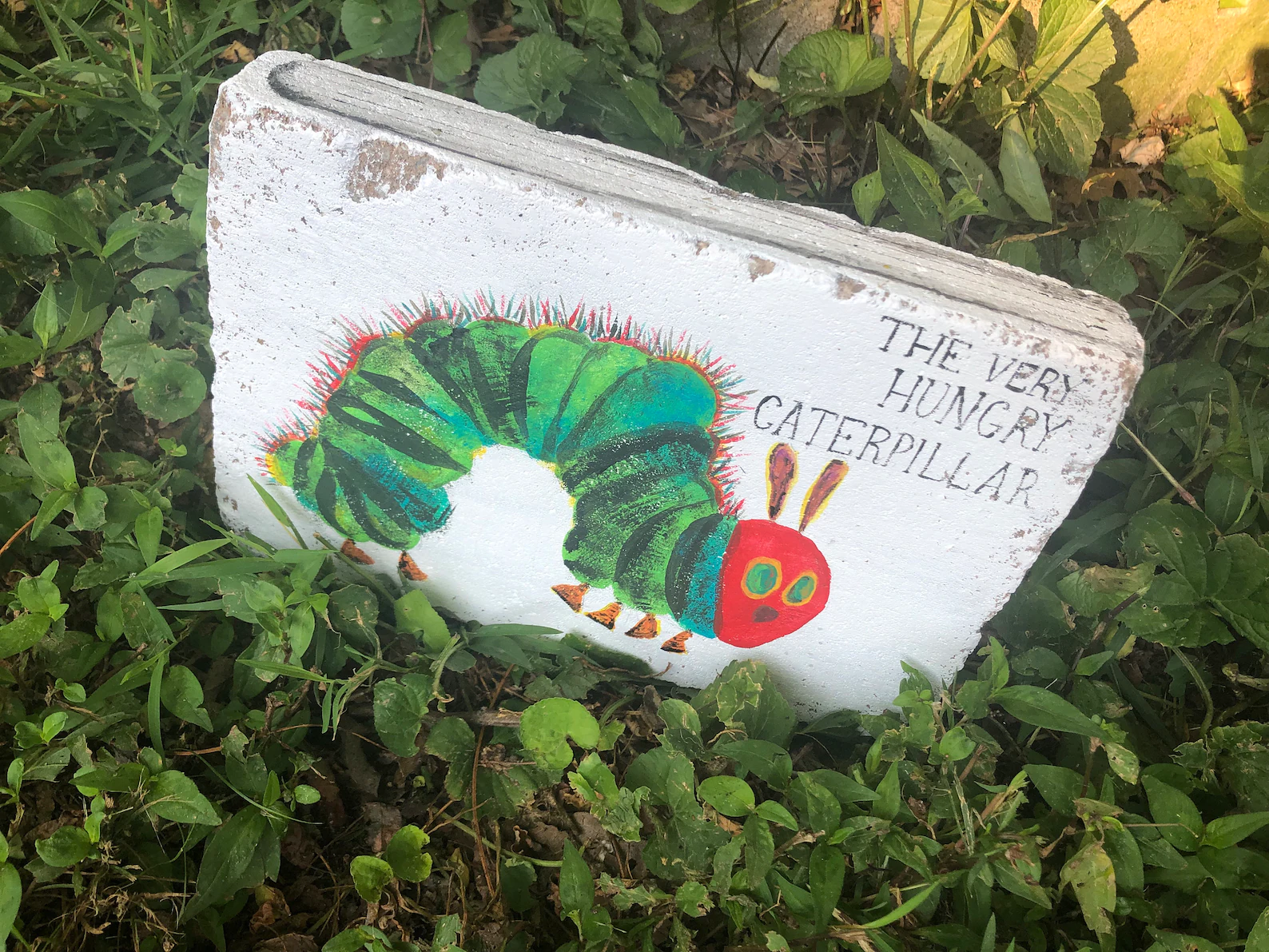A large flat stone painted to look like the cover of The Very Hungry Caterpillar sits in a small patch of garden.