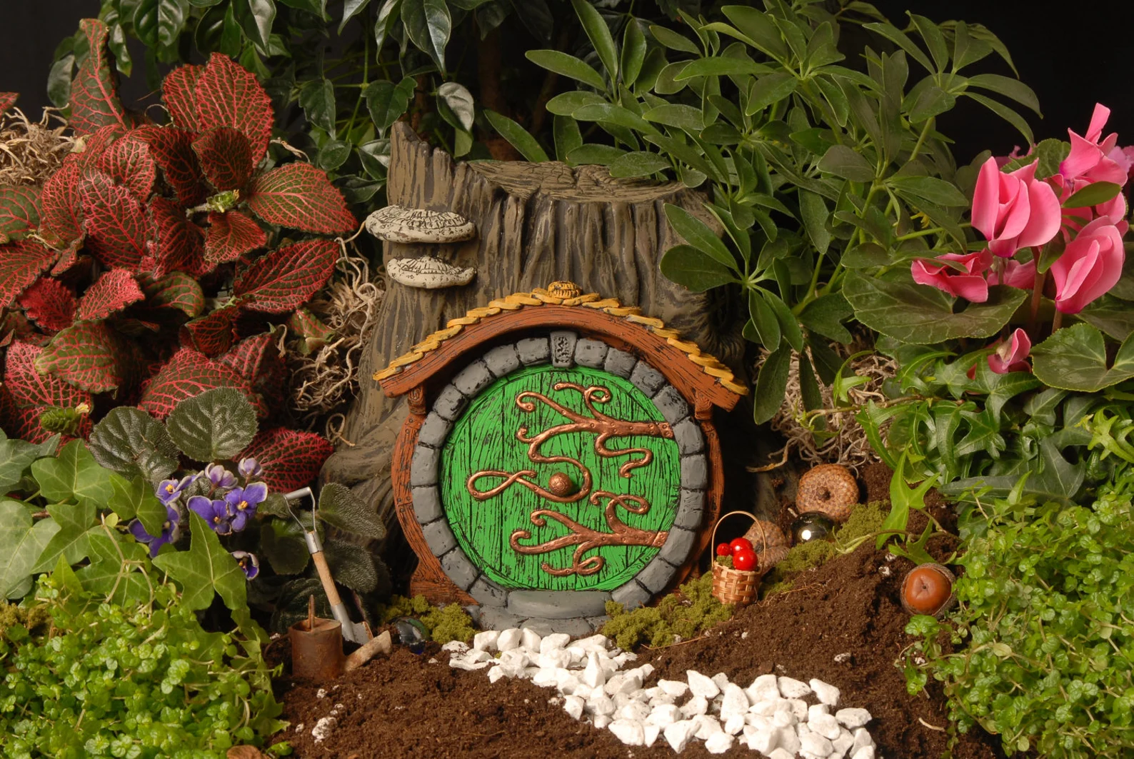 A small round green door is set at the base of a small stump, surrounded by plants, mini tools and baskets, and a tiny path made of small stones.