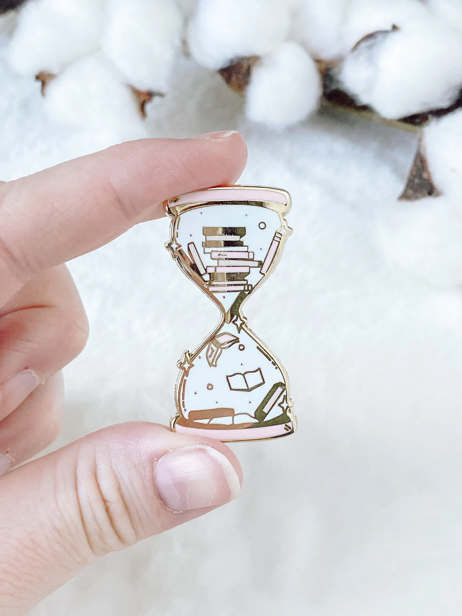 A pastel enamel pin depicting an hour glass with books inside