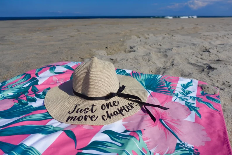 Image of a big floppy sunhat on a floral beach blanket on a beach. The hat reads "just one more chapter" in black. 