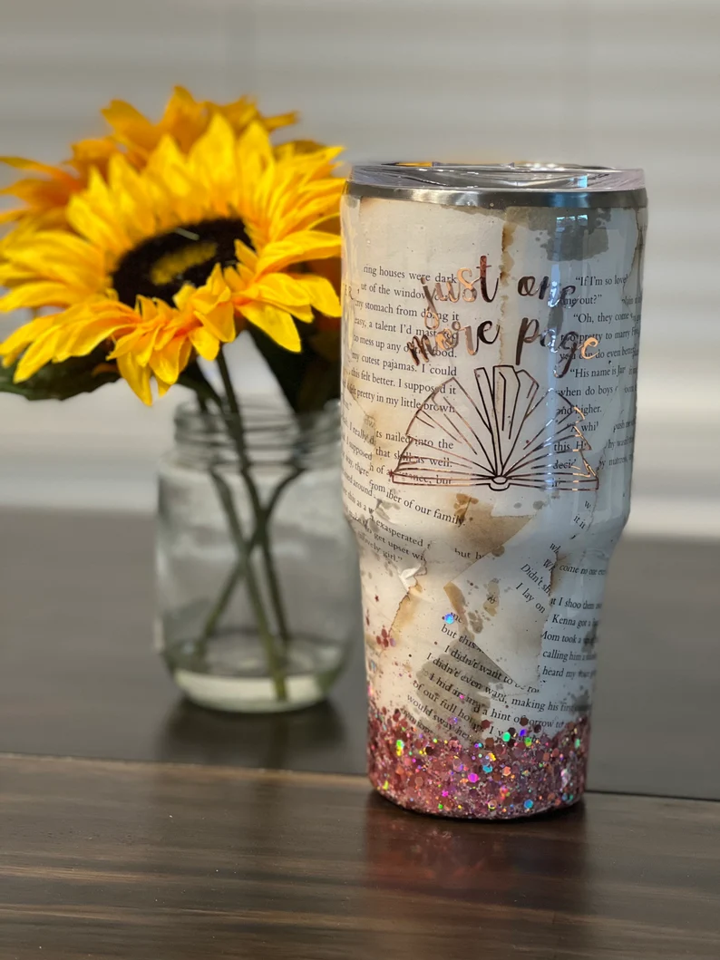 Image of a tumbler on a table. The tumbler is decorated with a book page, glitter, and the words "just one more page."