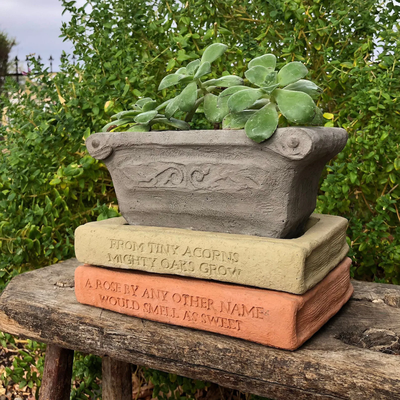 A cement planter consisting of two book-shaped saucers and a rectangular planter with a succulent plant sitting on top.  Quotations are engraved on the spine of the book.
