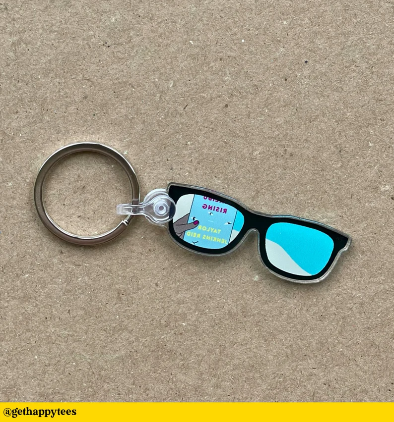 Image of a keychain in the shape of sunglasses. Reflected in one of the lenses is the book cover for Malibu Rising. 