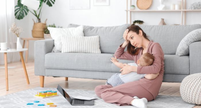 a tired mother nursing a child while looking at a laptop