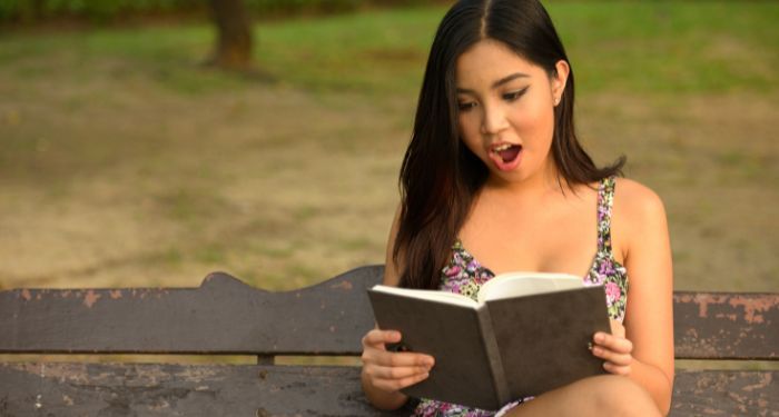 an Asian teen reading a book on a bench with a shocked expression