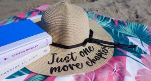 a photo of a sunhat with black script reading Just One More Chapter on a towel at the beach beside a stack of books