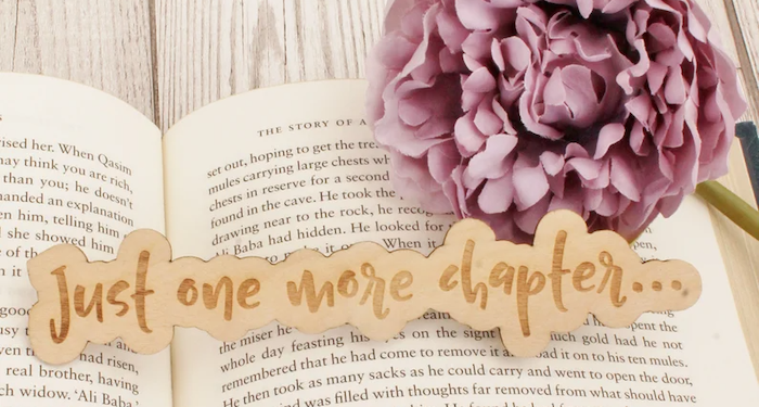 a photo of a wooden bookmark that says Just One More Chapter... resting on an open book beside a purple fabric flower