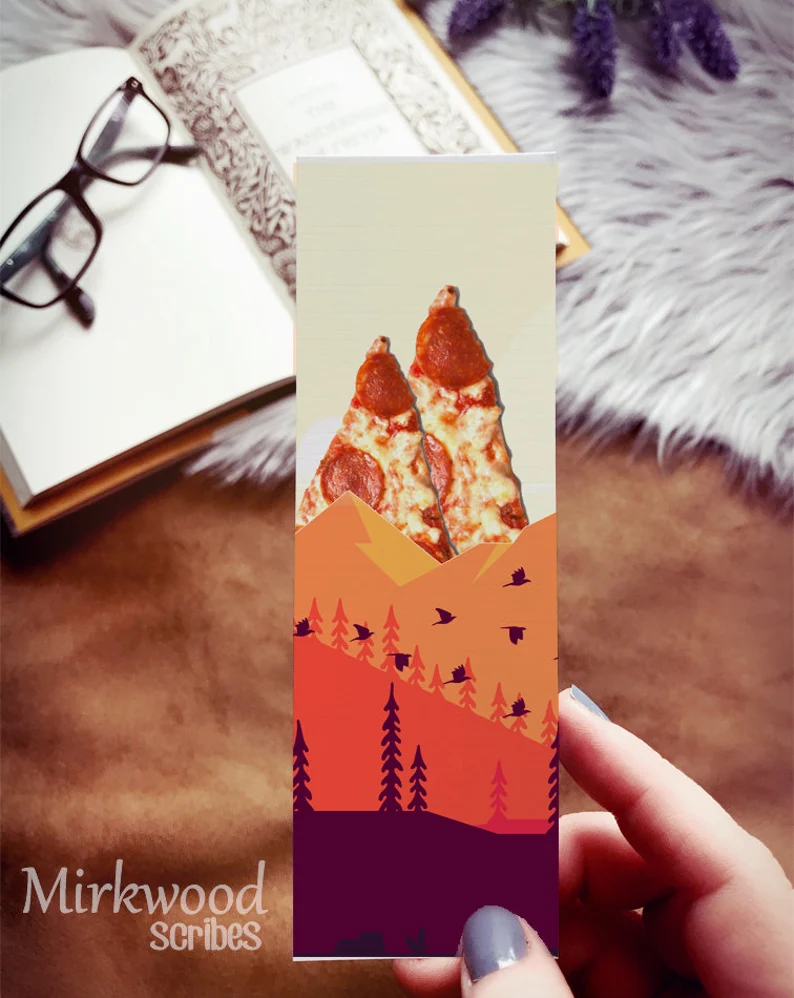 Image of a hand holding a bookmark of a mountain scene. The mountains in the back are pizza slices. 