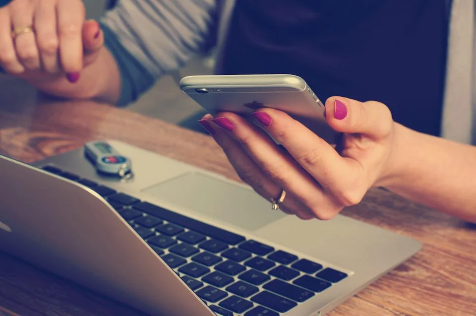 A person sits at a table in front of a laptop, with only their torso and hands visible in frame. They have red nail polish, and are holding a smartphone in their left hand.