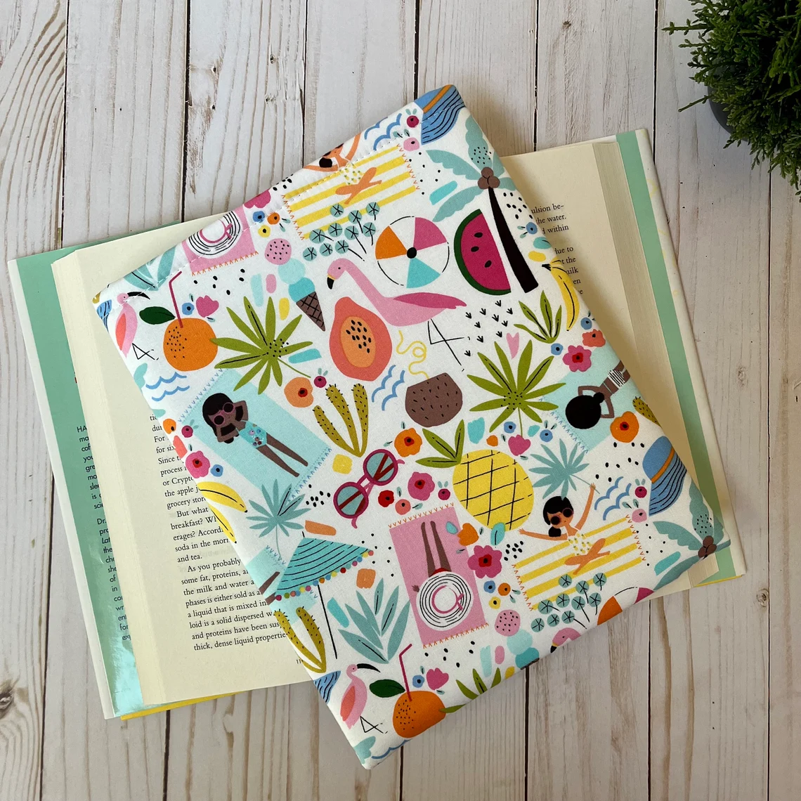 A brightly-colored book sleeve on top of an open book. The fabric of the book sleeve is summer themed, with beach blankets, beach bathers, and fruit. 