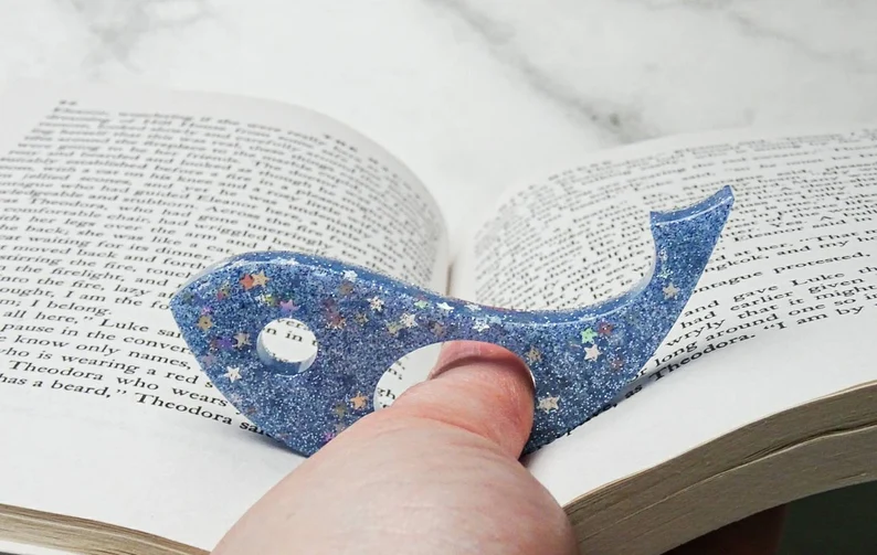 Image of a white hand holding a book page holder. The holder is blue and sparkly and shaped like a whale.