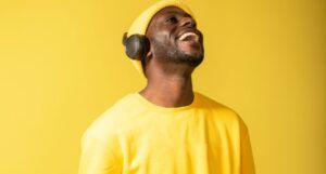 brown-skinned Black man smiling with black headphones, a yellow beanie, a yellow shirt, against a yellow background