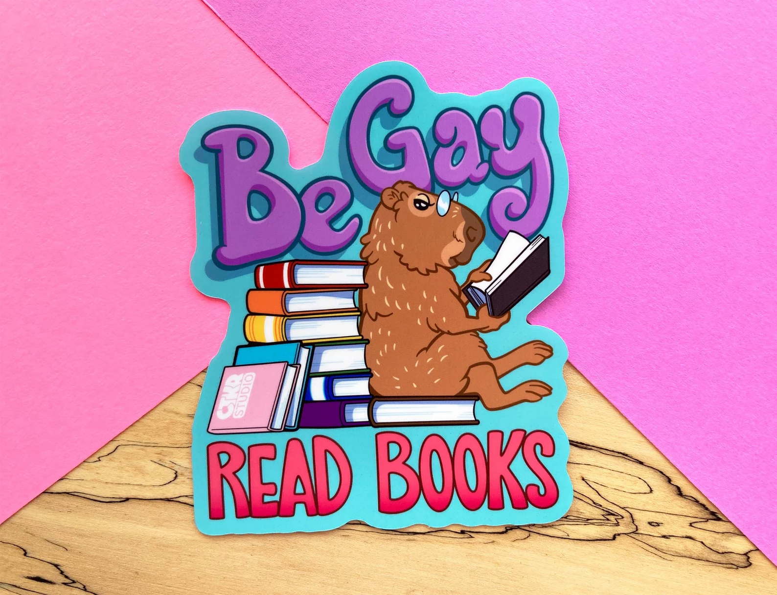 A colorful sticker that reads "be gay, read books" and depicts a reading capybara with a stack of rainbow colored books