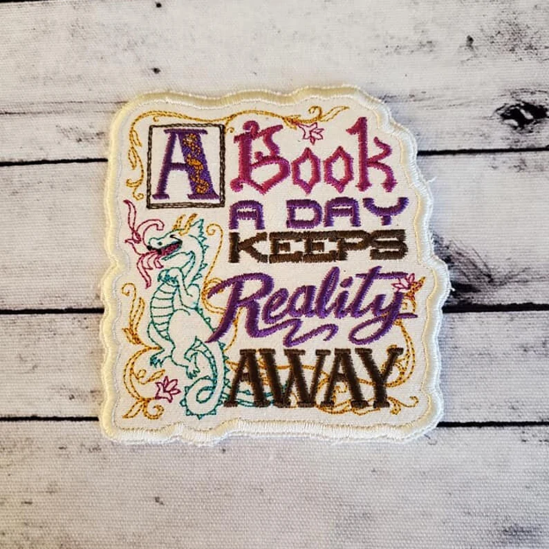 Image of an ecru patch that reads "a book a day keeps reality away." It has a green dragon image on it. 