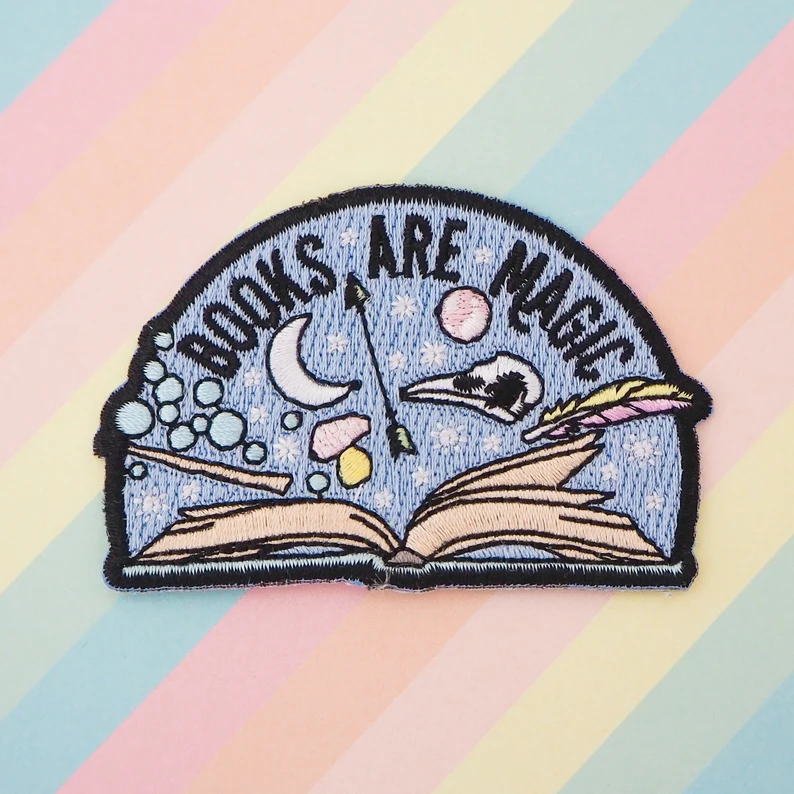 Image of a patch that features an open book. It has a moon, arrow, feather, and bird skull.. The patch reads "books are magic."