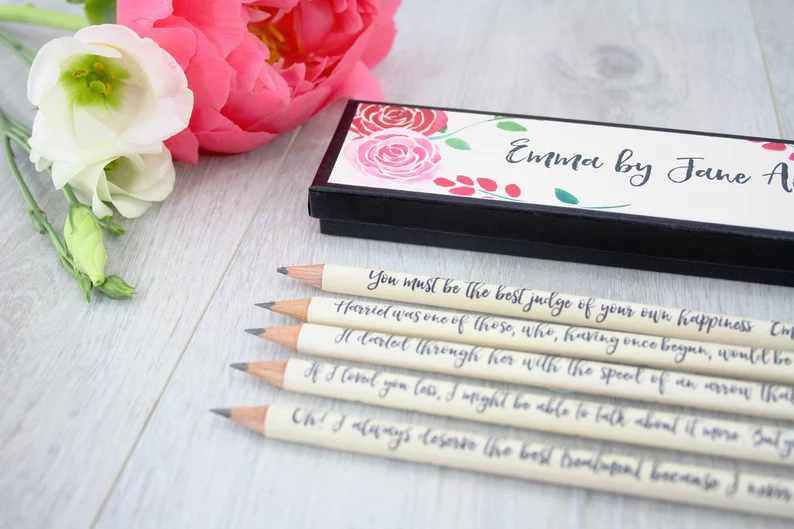 Photo of five pencils lined up showcasing Emma quotes, with what looks like a pencil case next to it, alongside some flowers