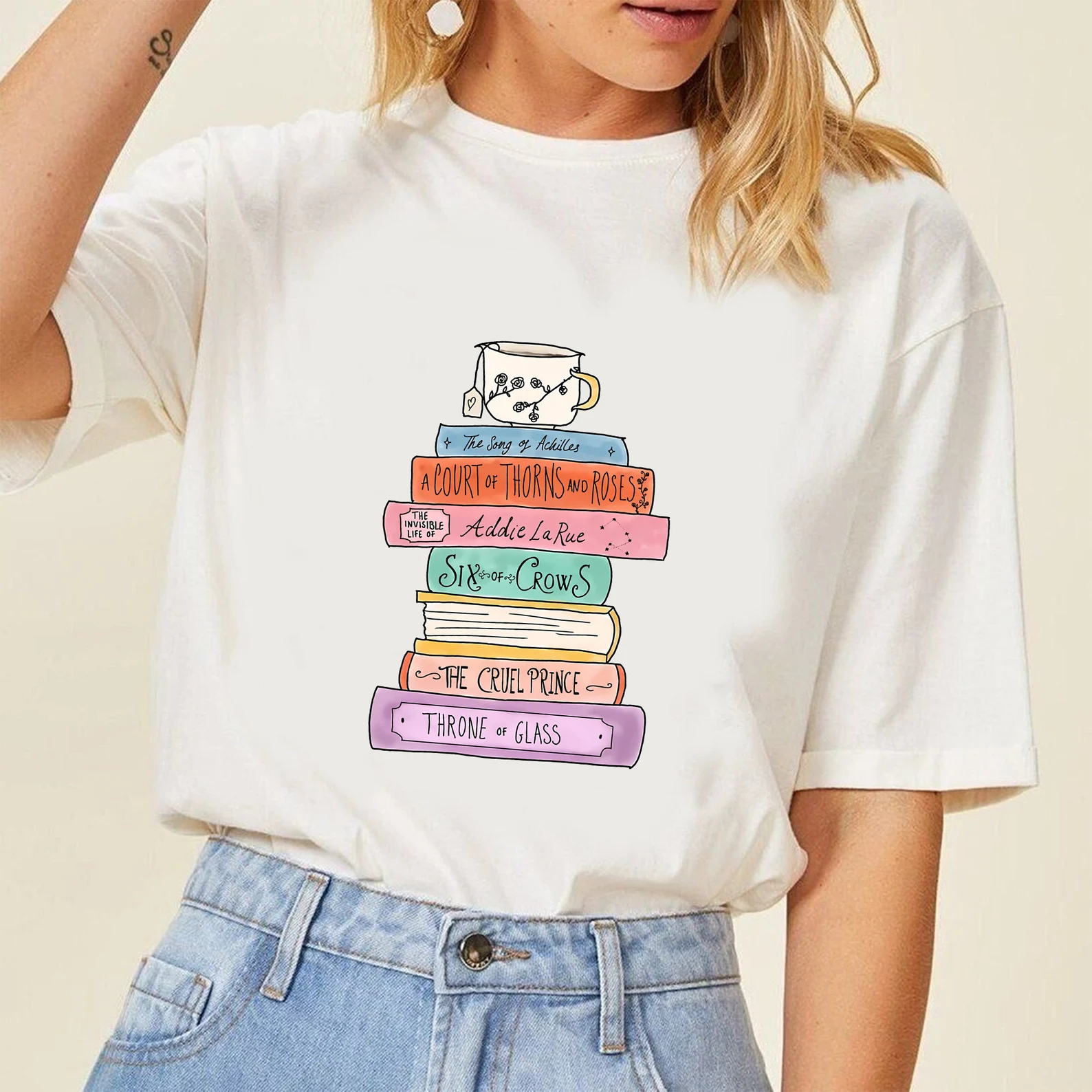 off-white t-shirt with a stack of pastel books whose spines display the titles Addie LaRue, The Cruel Prince, A Court of Thorns and Roses, Six of Crows, Throne of Glass, and Song of Achilles.