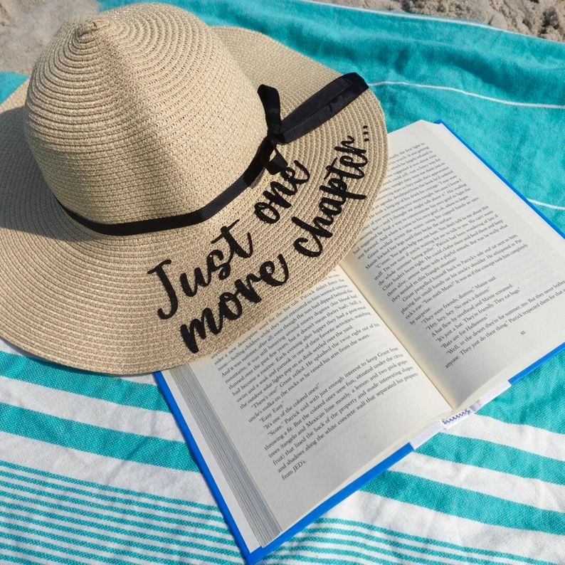Floppy hat with the words "just one more chapter" on a beach blanket. 