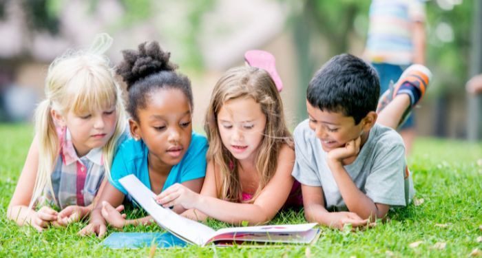 three girls and one boy of varying skin tones lie on the grass reading from the same book