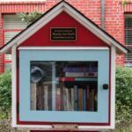 a red, white, and light blue Little Free Library in the shape of a house that is sitting in the yard of red brick house