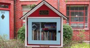 a red, white, and light blue Little Free Library in the shape of a house that is sitting in the yard of red brick house