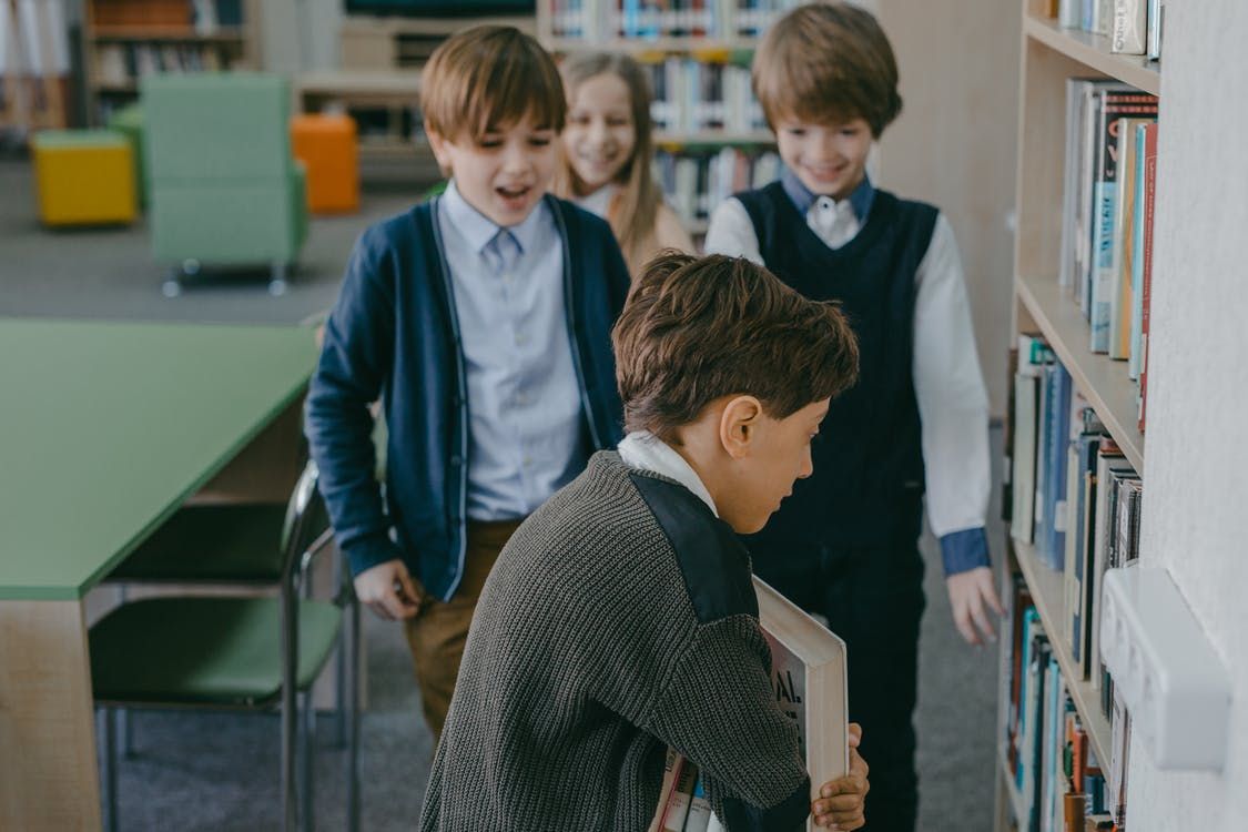 Four children are standing in a library. The child in the foreground is looking at books on a shelf, while the other three are watching and smiling. 